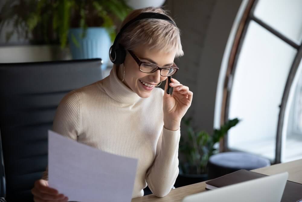 Smiling female worker employee wearing wireless earphones with mic, enjoying business negotiations video call meeting, discussing paper document, marketing research report with colleagues online.
